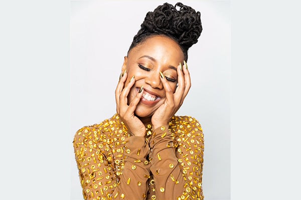 Headshot of a smiling woman in a tan, beaded top, cradling her face with both hands.