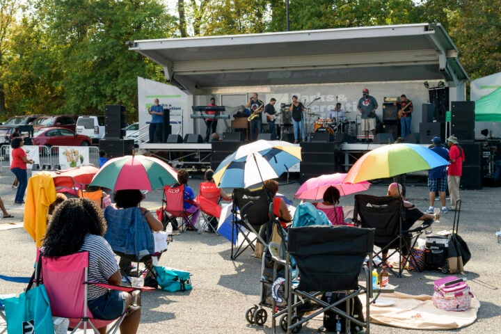 A band performs in a mobile bandwagon in a park, with audience members sitting in folding chairs on the pavement. Several use colorful umbrellas for shade.