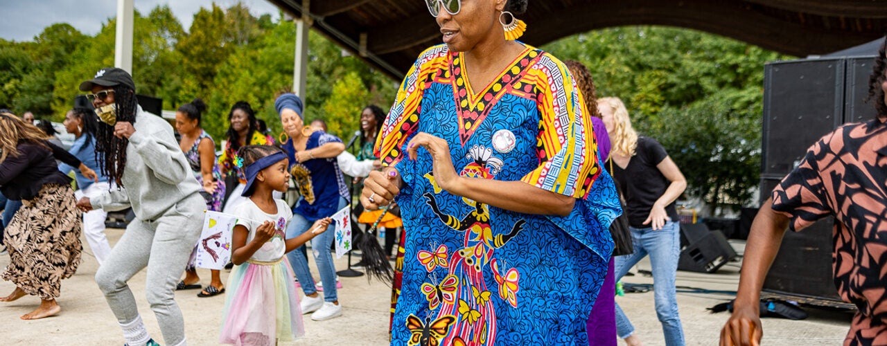 Festival attendees dance together in front of an outdoor stage, including a person in a bright blue patterned garment and a child in a pastel dress.