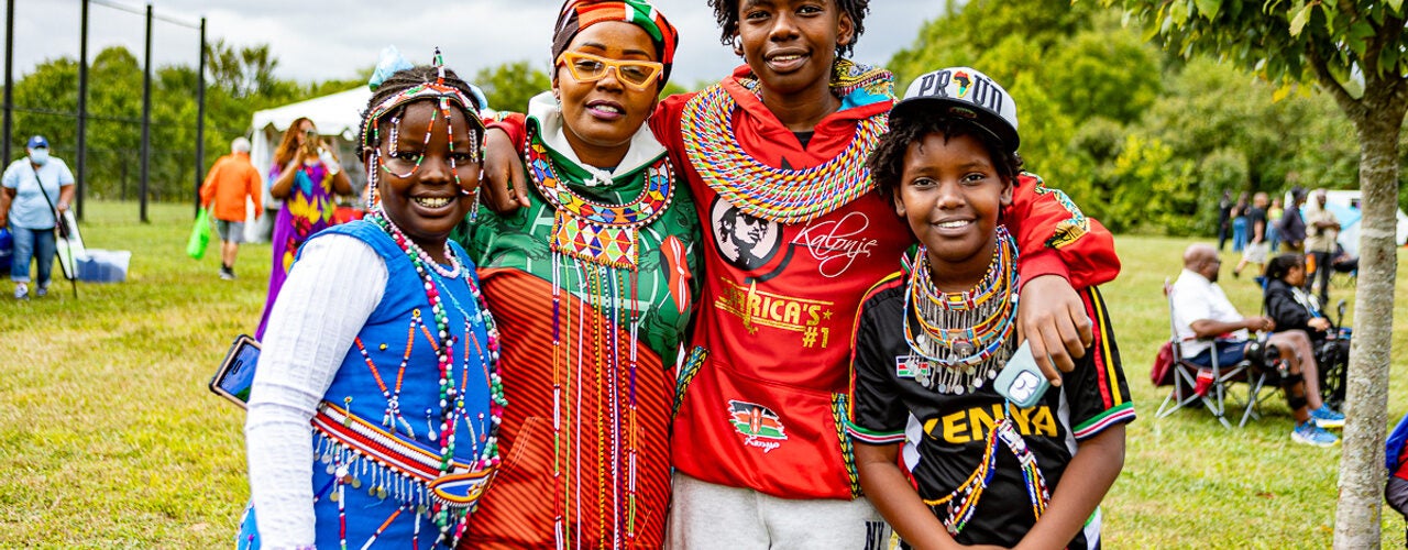 Four festival attendees pose outdoors wearing colorful beaded jewelry, patterned clothing, and accessories, with trees and event tents in the background.