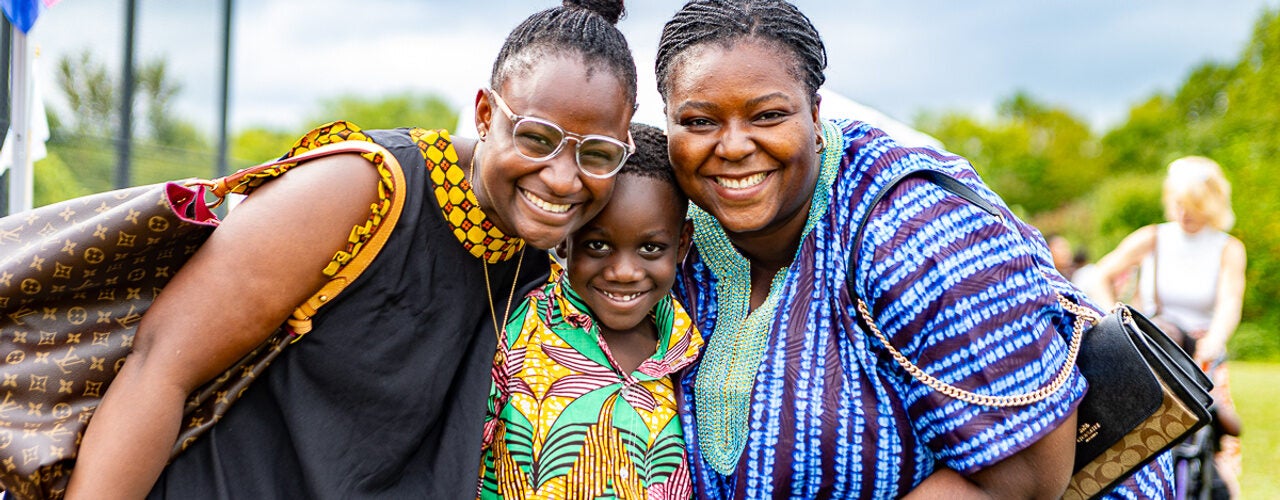 Three people pose closely together at an outdoor festival, wearing vibrant patterned clothing against a backdrop of grass, trees, and gathering attendees.
