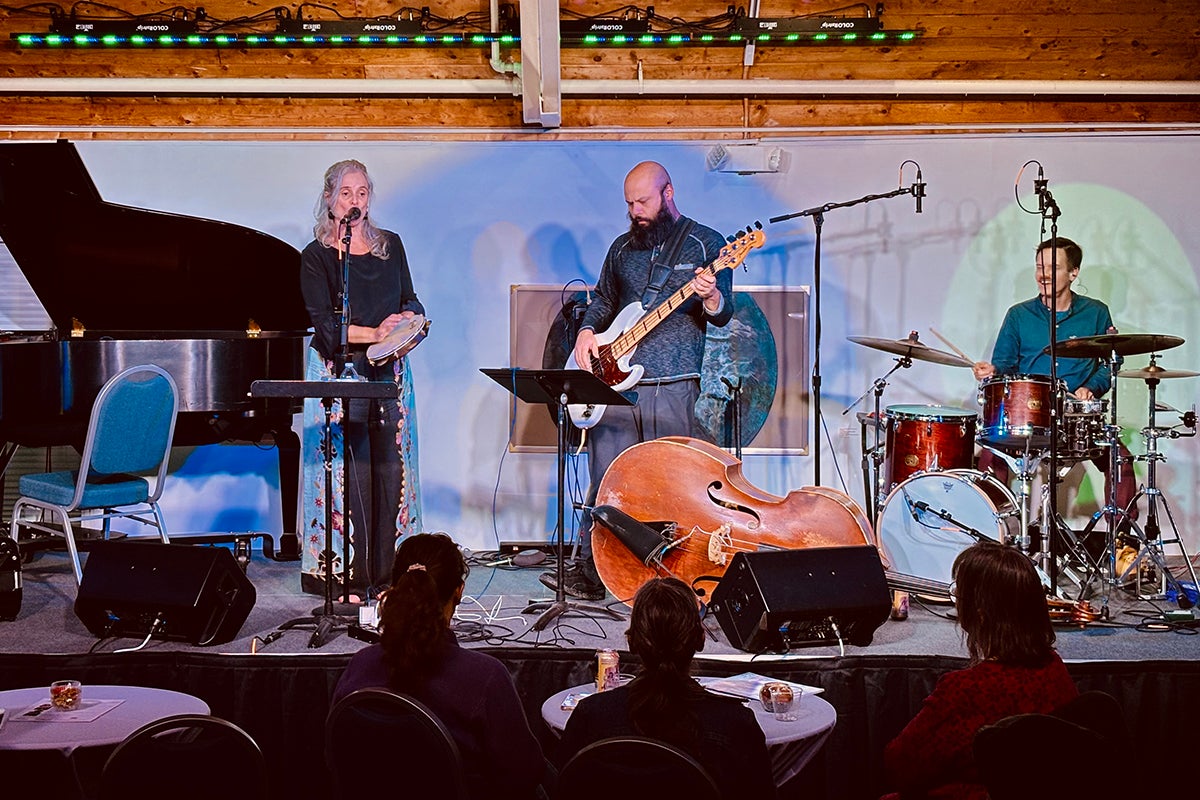 Three musicians perform on a stage in front of a seated audience. They include a singer/tambourine player, a drummer, and a bass guitarist.