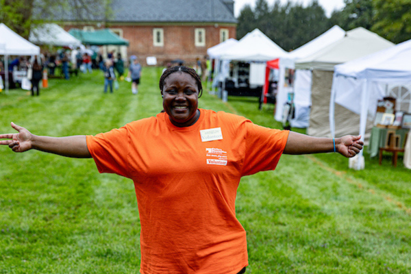 A smiling adult stands in the grass with arms outstretched. Behind her are two long rows of vendor tents.