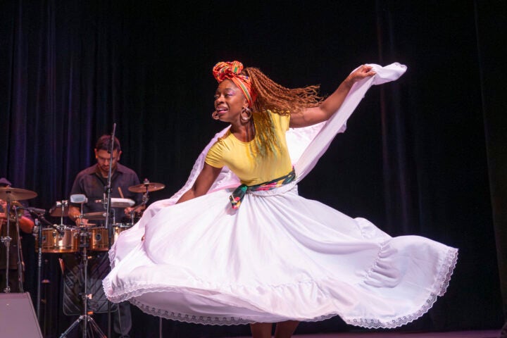 Performer in a yellow top and flowing white skirt twirls onstage beside live percussionists during a cultural dance performance.