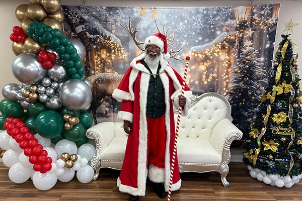 A man dressed in a Santa suit stands in front of wintertime photo backdrop and a white sofa positioned between two holiday trees.