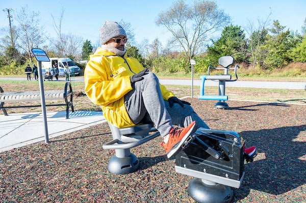 Person using a seated leg press machine at an outdoor fitness park, exercising on a rubberized surface with additional equipment in the background.
