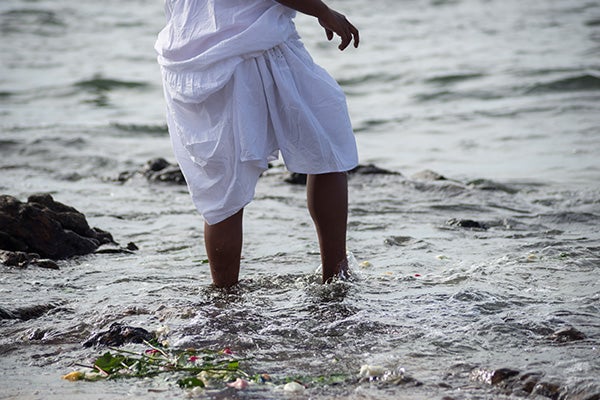 A person wearing white, wading into dark water. Flowers are washed up at the water's edge.