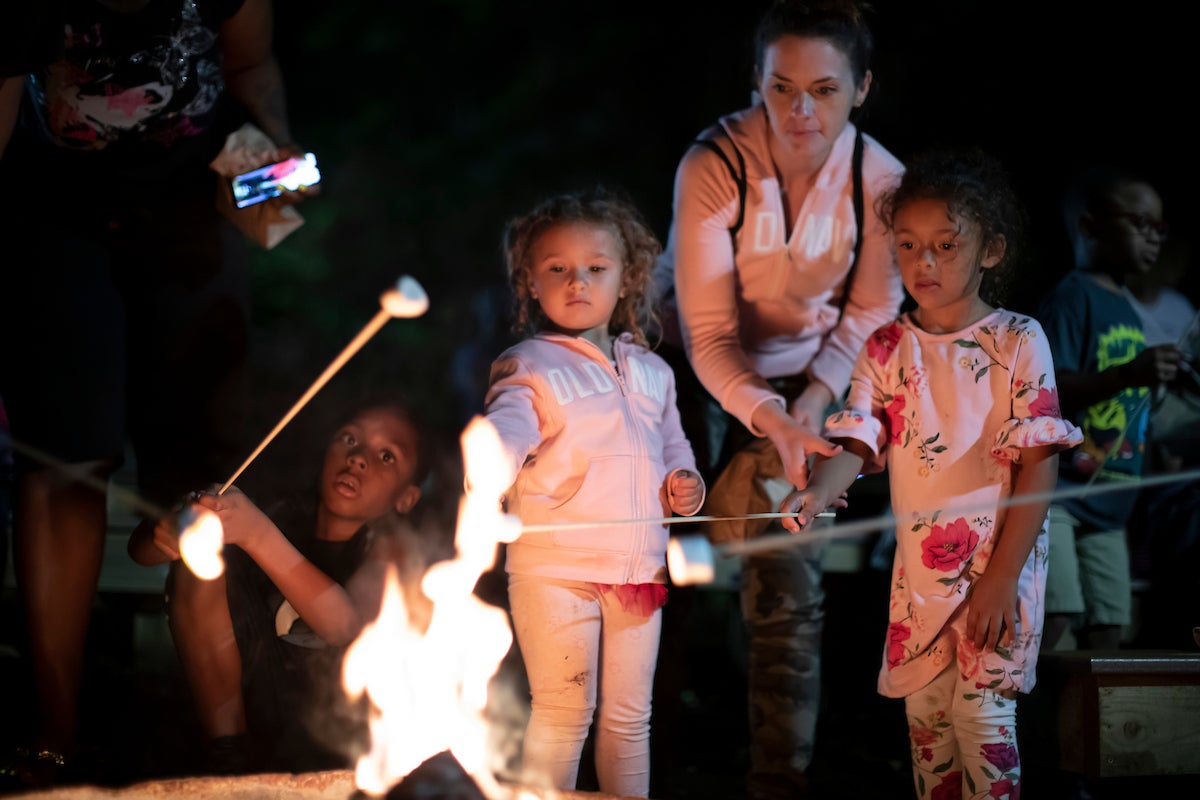 Parents supervise young kids as they toast marshmallows on long sticks over a campfire at night.