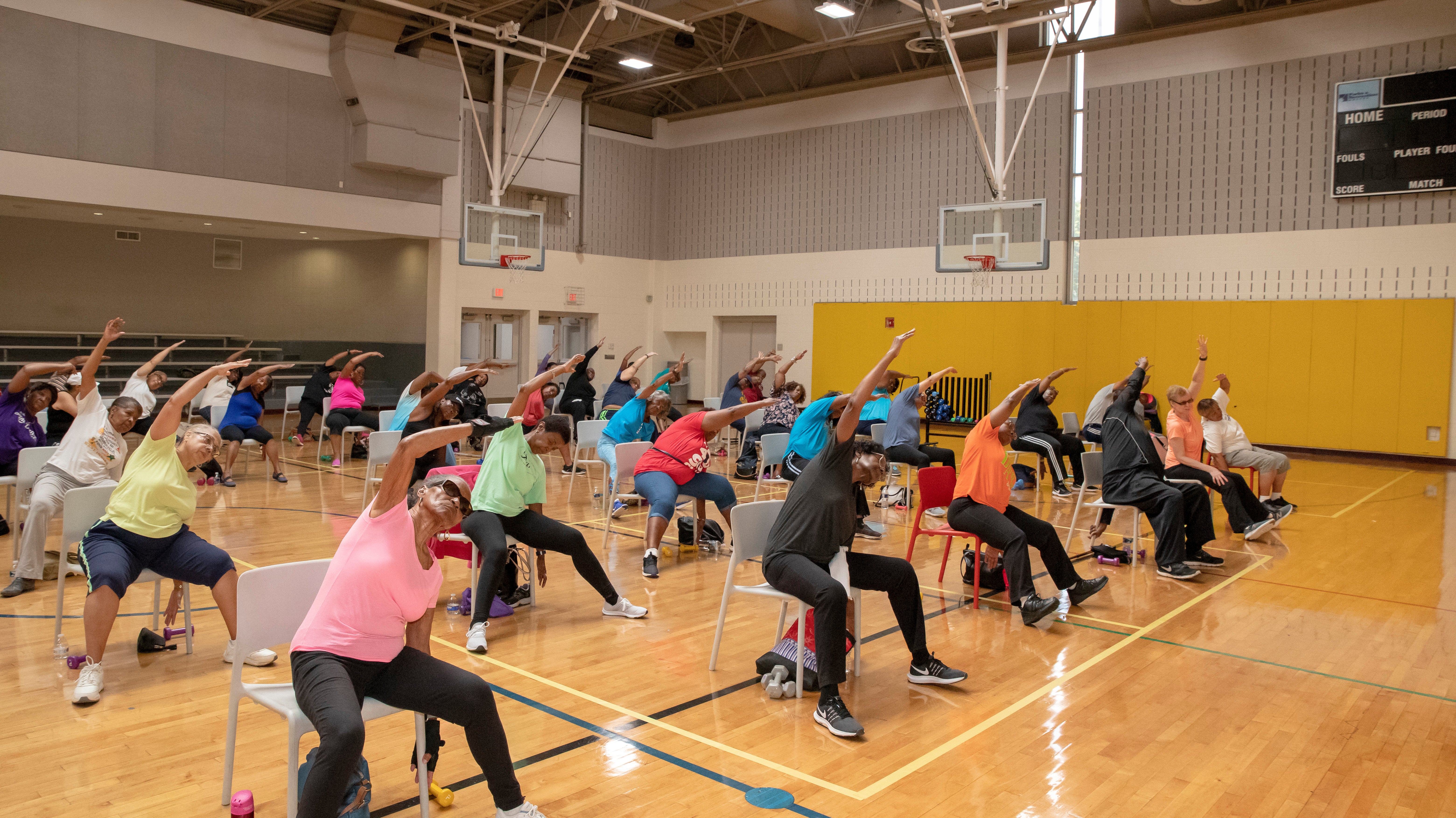 A large group of seniors performing side stretches while seated in chairs inside a gym.