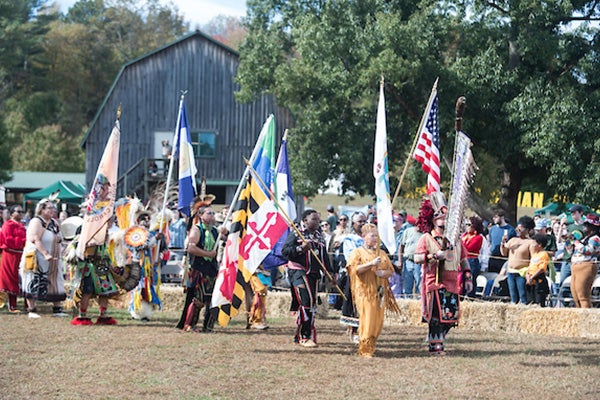 Spectators watch a parade procession at the American Indian Festival, led by people wearing native clothing and carrying flags.