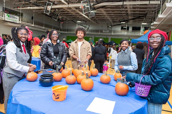 In a gym full of people, five smiling teens stand around a pumpkin-decorating table.