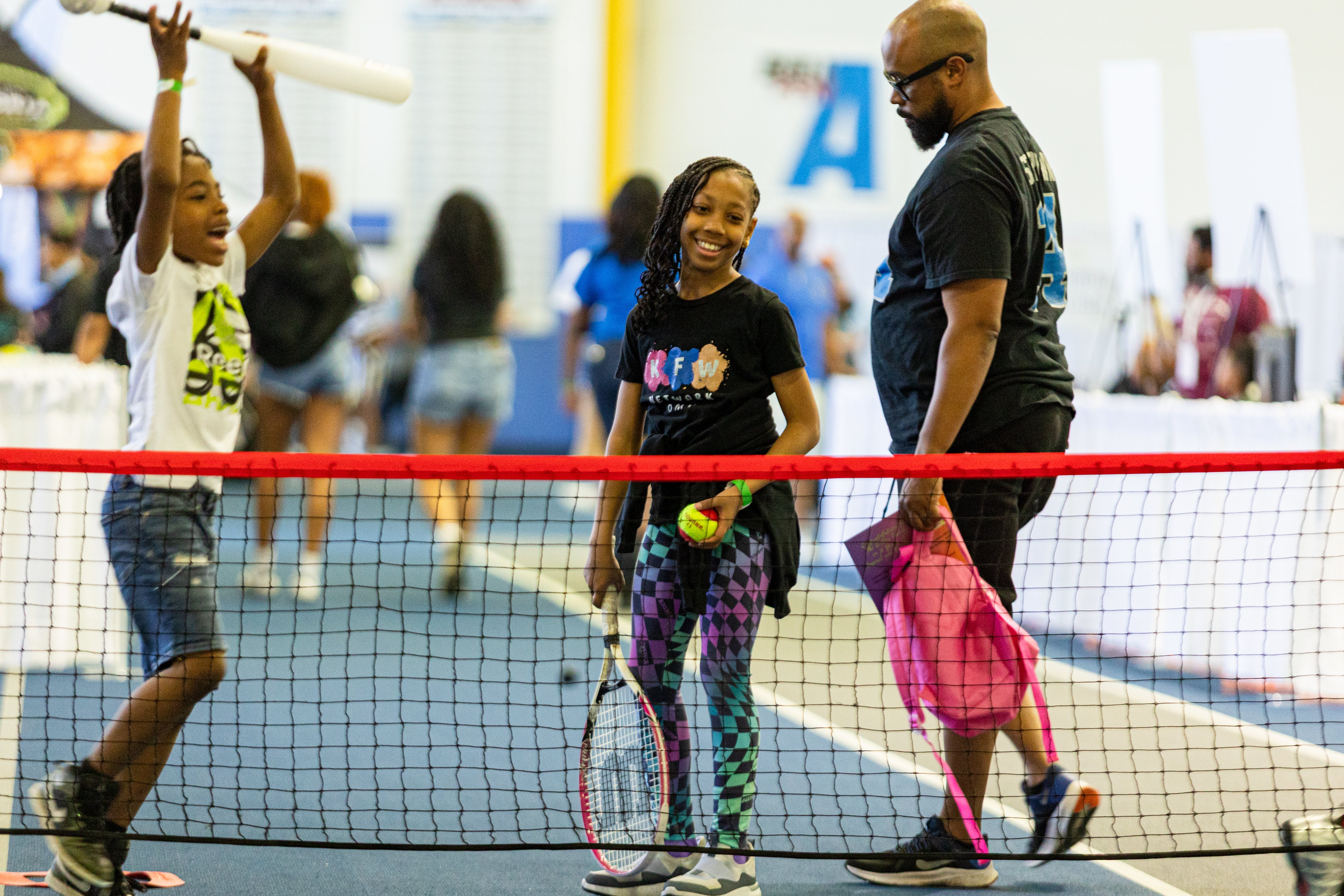 A girl holding a tennis racket and ball smiling near a net alongside a man and another playing child.